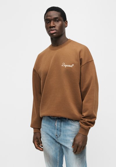 Νεαρός μαύρος άνδρας που φορά καφέ φούτερ Dsquared2 και ανοιχτό μπλε τζιν, στέκεται μπροστά σε απλό φωτεινό φόντο.