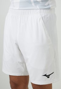 Weiße Sportshorts mit elastischem Bund, seitlichen Taschen und einem schwarzen Logo am unteren linken Bein, ausgestattet mit einer glatten Textur.
