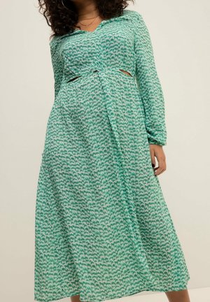Femme portant une longue robe verte et blanche à motifs, manches longues et petites découpes latérales à la taille, debout devant un fond uni.