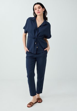 Vrouw staat met de handen in de zakken, draagt een marineblauwe blouse met knopen en trekkoord in de taille, bijpassende broek en bruine sandalen tegen een egale achtergrond.