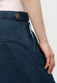 Λεπτό χέρι ακουμπά δίπλα σε σκούρο μπλε τζιν φούστα με ψηλή μέση, με καφέ κουμπί και ορατή ραφή πλαϊνής τσέπης.