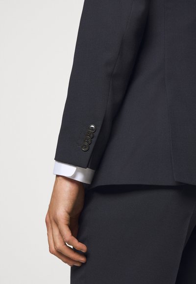 Casaco de fato escuro, ajustado, com tecido texturizado, apresentando quatro botões pretos na manga e um punho de camisa branca visível por baixo.