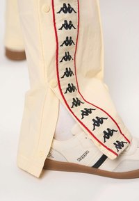 Bézs színű melegítőnadrág fekete és piros logószalaggal az oldalán, valamint egy fehér Kappa sneaker gumitalppal és fekete csíkkal.