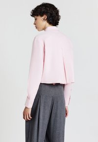 Person mit kurzem, lockigem Haar, die ein hellrosa, kurzes Langarmshirt und eine hoch taillierte, graue, gestreifte, weite Hose trägt, von hinten betrachtet.