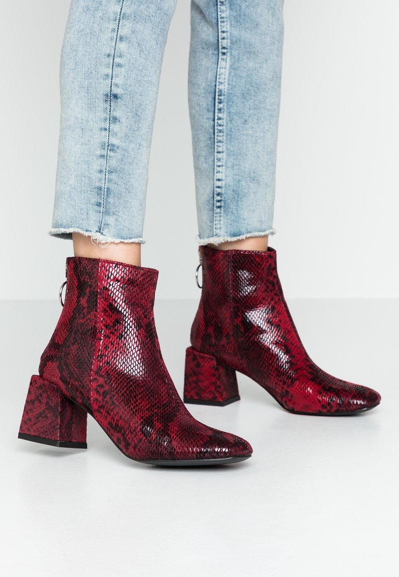 Bottes cheville en cuir de serpent rouge avec une finition brillante, talons carrés et un détail zippé. Le look est complété par un jean en denim à ourlet effiloché.