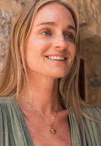 Gold necklace with sunburst pendant and a pearl accent. The model has long blonde hair and wears a green textured garment.