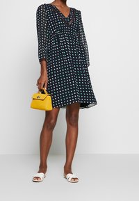 Kvinna klädd i en marinblå klänning med prickar, håller en liten gul handväska, kombinerad med vita sandaletter, stående på ett vitt golv.