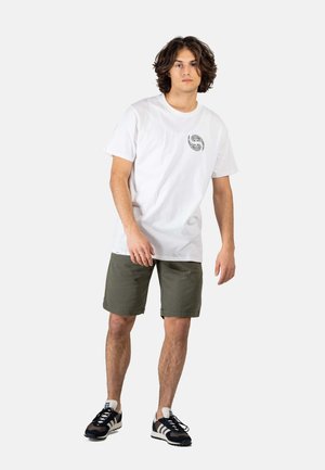 Joven de pie con una camiseta blanca con gráfico, pantalones cortos verde oliva y zapatillas deportivas blancas y negras sobre un fondo liso.