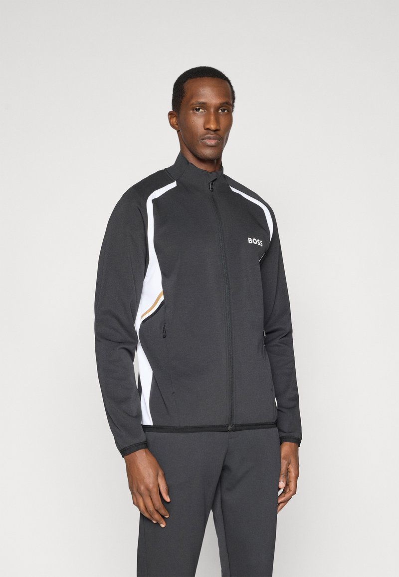 Casaco preto com fecho de correr e detalhes em branco e dourado. Apresenta um colarinho alto, bolsos laterais e um design desportivo. Feito de tecido suave.