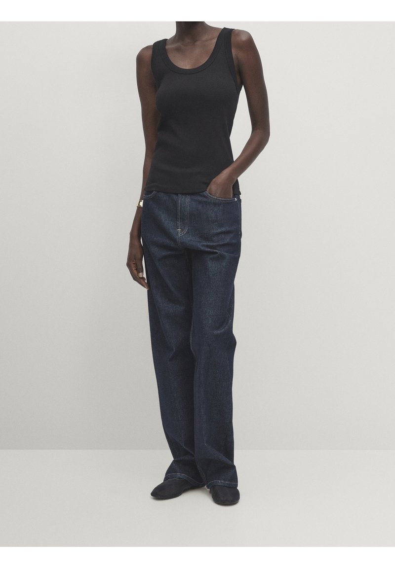 Femme portant un haut noir sans manches, un jean bleu foncé ample et des chaussures noires, debout avec une main dans la poche devant un fond uni.