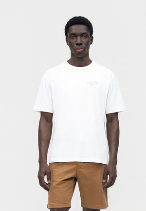 Mladý muž stojí čelom dopredu, oblečený v biele tričko s krátkym rukávom s logom a hnedé šortky po kolená na jednoliatom pozadí.