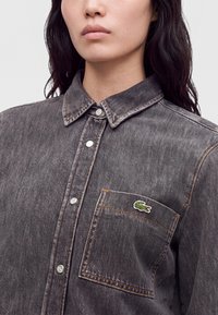Gros plan d'une personne portant une chemise en denim gris foncé avec des boutons-pression métalliques et une poche poitrine ornée d'un logo crocodile vert.