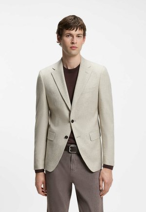 Junger Mann, der einen hellbeigen Blazer über einem dunkelbraunen Hemd mit grauer Hose und schwarzem Gürtel trägt, vor einem schlichten weißen Hintergrund stehend.