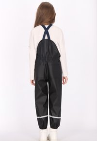 Vaikas, vilkintis juodas nepralaidžias kelnes su tamsiai mėlynomis juostelėmis, kurios kryžiuojasi nugaroje, baltą ilgomis rankovėmis marškinį ir baltas batus, stovi atsukęs nugarą.