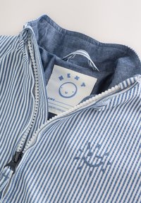 Vêtement zippé à rayures bleues et blanches avec une broderie de soleil souriant et une étiquette de marque "NEXT" à l'intérieur du col.
