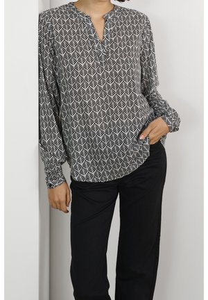 Frau trägt eine schwarz-weiß gemusterte Langarmbluse mit Blättermotiv und schwarze Hose, steht mit einer Hand in der Tasche vor weißem Hintergrund.