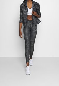 Mujer con chaqueta negra con estampado y leggings, que lleva un sujetador deportivo blanco y zapatillas blancas, de pie contra un fondo claro y liso.