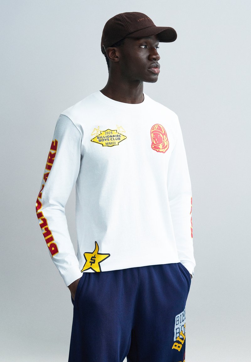 Λευκό πουκάμισο με μακριά μανίκια  με κόκκινες και κίτρινες γραφικές εκτυπώσεις. Διαθέτει αστέρι με σύμβολο του δολαρίου και κείμενο κατά μήκος των μανικιών. Σκούρο παντελόνι.