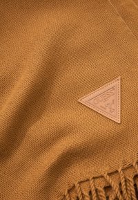 Brązowa tkanina o fakturze, z frędzlami na krawędziach, ozdobiona trójkątną beżową skórzaną naszywką z wytłoczonym napisem "GUESS".