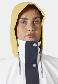 Giacca impermeabile bianca con cappuccio giallo. Presenta una patta blu navy con un bottone a pressione argento, un cordino regolabile e una texture liscia.