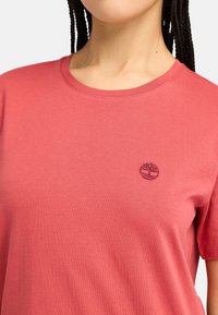 T-shirt en coton rouge avec un col rond, des manches courtes et un petit logo brodé sur la poitrine. Texture douce et coupe décontractée.