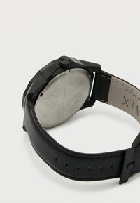 Črna ura z ostrimi linijami. Ima jekleno hrbtno plat, gladek črn usnjen pašček in okroglo analogno številčnico.