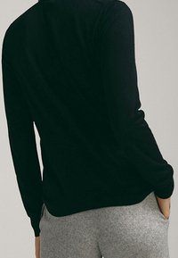 Maglione nero a maniche lunghe con lavorazione a maglia testurizzata, stile aderente e polsini a costine. La vista dal retro mostra una silhouette liscia. Abbinato a pantaloni grigi.