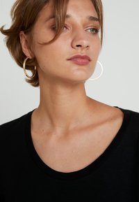 T-shirt preta com um decote amplo, complementada por grandes brincos de argola dourados. O tecido parece suave, com um brilho sutil e um corte descontraído.