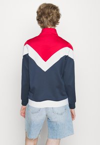 Giacca color block in blu navy, rosso e bianco con colletto alto e design a chevron. Presenta una vita testurizzata con dettagli del logo.