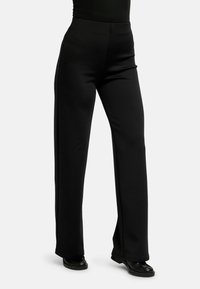 Žena oblečená v čiernych širokých nohaviciach s vysokým pásom v kombinácii s čiernymi topánkami na jednoduchom svetlom pozadí.