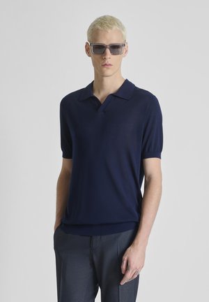 Joven con pelo rubio corto que lleva gafas de sol oscuras, camisa tipo polo de manga corta azul marino y pantalones oscuros, de pie frente a un fondo liso.