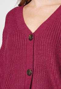 Fuchsia knit cardigan with a deep V-neck and two brown buttons. Features ribbed texture, long sleeves, and a relaxed fit.
