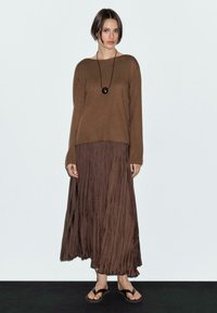 Brauner gestrickter Pullover, langer brauner Plisseerock, braune flache Sandalen und eine schwarze Anhänger-Halskette. Neutrale Hintergrundgestaltung, minimalistisches Design.