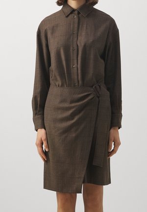 Kvinde iført brun skjortekjole med lange ærmer og matchende bælte, der er fastgjort i siden, stående mod en ensfarvet baggrund.