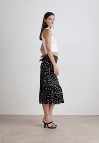 Falda midi negra con estampado floral y dobladillo rizado, fabricada en un tejido ligero, combinada con una camiseta de tirantes blanca ajustada y sandalias negras de tacón.