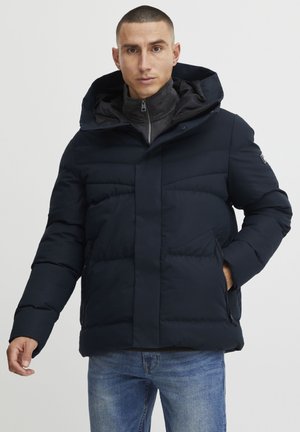 Navy puffer jacket with a hood, featuring a zip closure, side pockets, and a quilted design. Worn over a gray high-collar sweater.