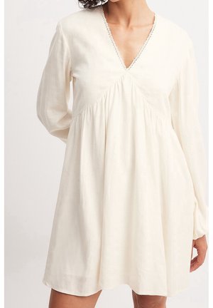 Biała sukienka z długim rękawem, dekoltem w szpic i marszczoną talią, noszona przez osobę o kręconych włosach, stojącą z jedną ręką za plecami.