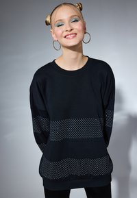 Felpa nera con maniche ampie, caratterizzata da righe orizzontali di piccoli punti bianchi. Tessuto morbido con una vestibilità rilassata e scollo a girocollo.