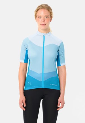 Frau mit lockigem blondem Haar, die ein hellblau-türkisfarbenes Radtrikot mit kurzen Ärmeln und schwarze Radhosen trägt, vor weißem Hintergrund stehend.