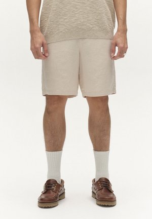 Man draagt beige gebreide korte broek, een beige gestructureerde top, witte enkelsokken en bruine leren mocassins, staande tegen een witte achtergrond.