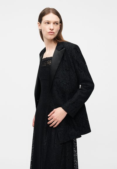 Γυναίκα που φορά ένα μαύρο φόρεμα χωρίς τιράντες από δαντέλα και ταιριαστό μαύρο σακάκι από δαντέλα με σατέν πέτα, κοιτάζοντας προς τα πλάγια σε απλό φόντο.