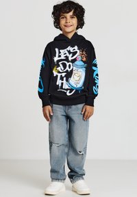 Felpa nera con design di graffiti colorati, che presenta il testo "FACCIAMO QUESTO." Indossata con jeans larghi di lavaggio chiaro e sneakers bianche.