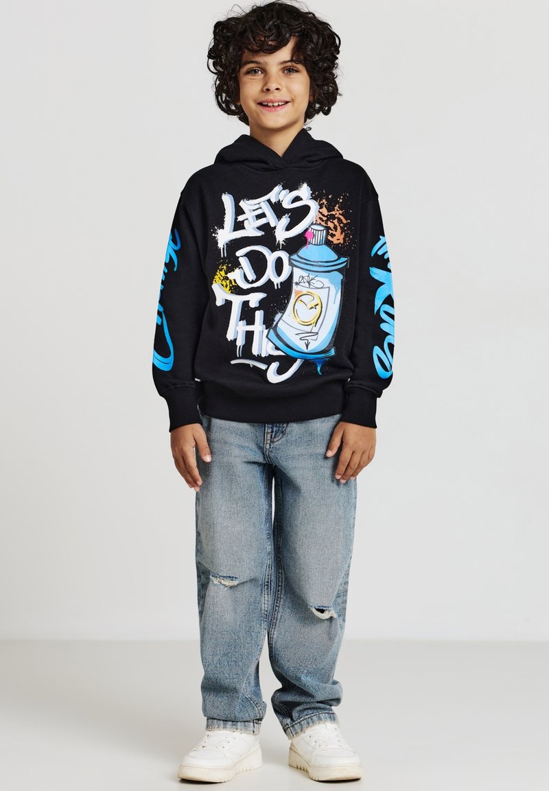 Felpa nera con design di graffiti colorati, che presenta il testo "FACCIAMO QUESTO." Indossata con jeans larghi di lavaggio chiaro e sneakers bianche.