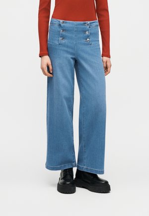 Nő magasderekú, bő szárú kék farmernadrágot visel, amelynek hat elülső gombja és nagy zsebei vannak, fekete vaskos bakancsokkal és rozsdaszínű felsővel párosítva.