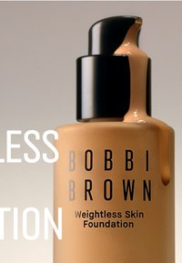 Bobbi Brown Weightless Skin Foundation i en klar glassflaske med et svart pumpehette. Væsken er i en middels brun nyanse med en glanset finish.