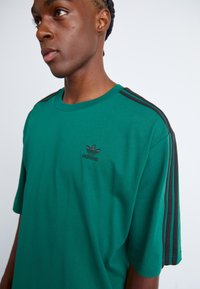Grøn kortærmet t-shirt med sort tre-stribe design på skuldrene. Har et broderet sort Adidas-logo på brystet.