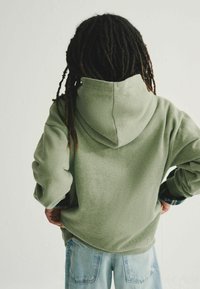 Sudadera verde con una textura suave y una capucha grande, combinada con jeans de mezclilla azul claro. Los puños presentan un acento de cuadros.