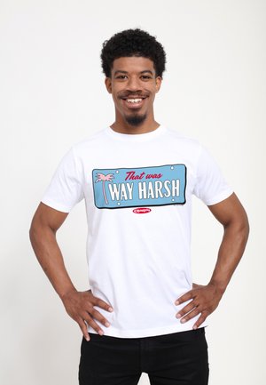 Lächelnder Mann mit lockigem Haar, der ein weißes T-Shirt mit der Aufschrift „Das war SEHR HART“ und einer pinken Palmen-Grafik trägt.
