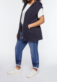 Nainen, joka on pukeutunut valkoisiin tennareihin, löysiin sinisiin farkkuihin, valkoiseen T-paitaan ja ilmanhihaiseen tummansiniiseen vetoketjulliseen huppariin, jolla on käsi taskussa.