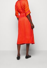 Röd klänning med geometriska mönster, knälängd, korta ärmar. Tyget verkar lättviktigt med en texturerad yta. Knuten i midjan.